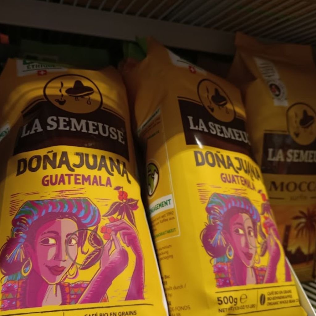 Café orgánico en grano “Doña Juana Guatemala” de la marca suiza La Semeuse, con su característico empaque amarillo e ilustración de una mujer recolectora de café, expuesto en un estante de supermercado.