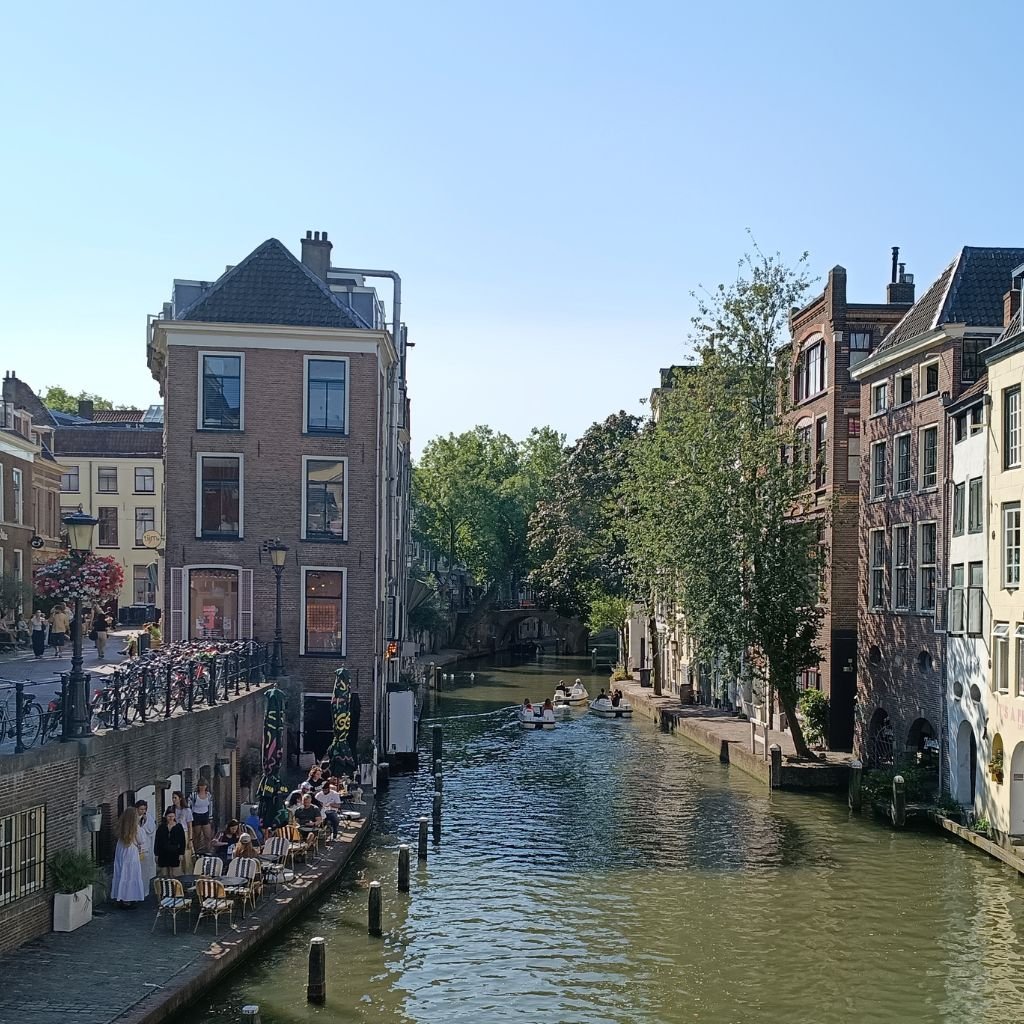 Terraza soleada junto al canal de Utrecht donde locales y visitantes disfrutan café al aire libre entre edificios históricos y barcas.